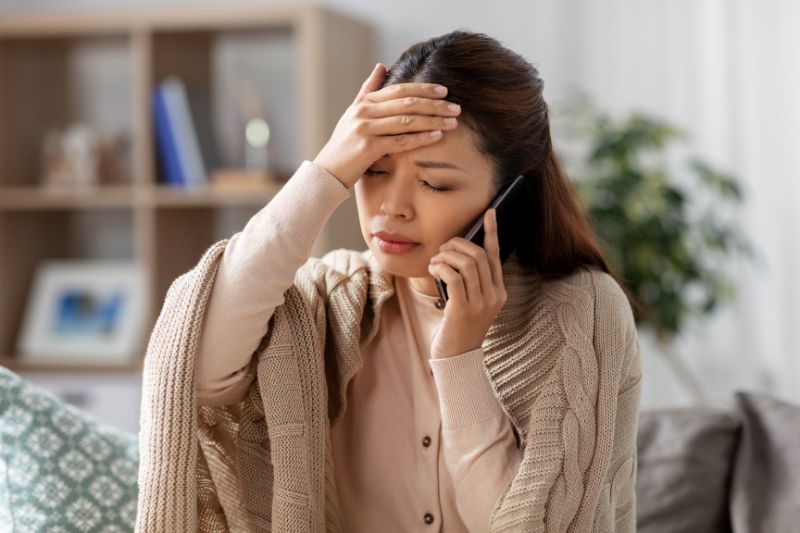 Dark haired lady looking very unwell, wrapped in an oatmeal textured wool blanket, holding her right hand to her forehead and her left hand holding a mobile ohone
